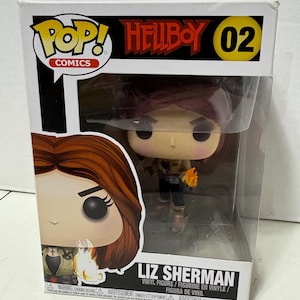 May include: A boxed Liz Sherman vinyl figure from the Hellboy series. The figure is in a clear plastic display case. The box features the Pop! Comics logo, the Hellboy logo, and the number 02. The figure is holding a flame.