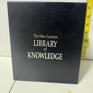 Peut inclure: Une boîte noire avec le texte "The New Lexicon LIBRARY of KNOWLEDGE" en blanc. Un ruban à mesurer jaune est visible sur le côté droit de la boîte, avec des mesures en centimètres.