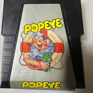 Peut inclure: Une cartouche de jeu NES vintage pour Popeye, avec une image de dessin animé de Popeye tenant une boîte de spinach et une bouée de sauvetage. La cartouche est noire avec une étiquette grise.