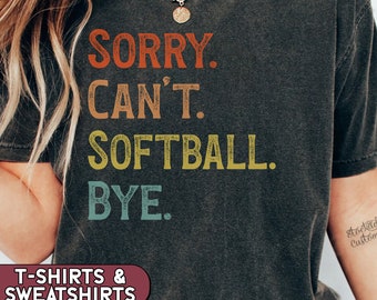 May include: A black t-shirt with a vintage-style graphic that reads "SORRY. CAN'T. SOFTBALL. BYE." The text is in rainbow colours. The t-shirt is available in a variety of sizes and styles.