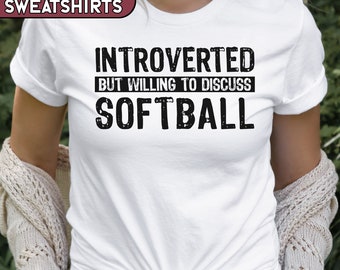 May include: White t-shirt with black text that reads "Introverted but willing to discuss Softball".