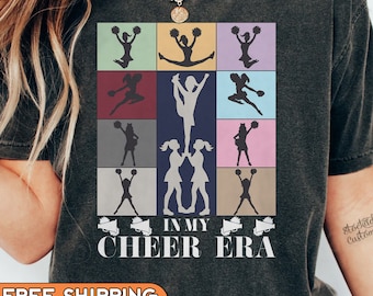 Könnte beinhalten: Ein dunkelgraues T-Shirt mit einer Grafik im Vintage-Stil von Cheerleadern in verschiedenen Posen. Die Grafik ist von einem bunten Rand umgeben und enthält den Text "In My Cheer Era".