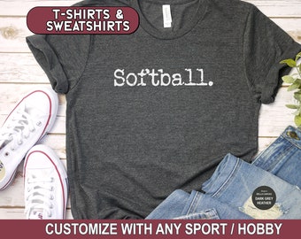May include: A dark grey t-shirt with the text "Softball." printed in white. The t-shirt is laid flat on a wooden surface with a pair of white sneakers and a pair of blue jeans.