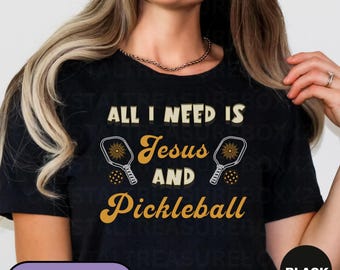 May include: Black t-shirt featuring the text "ALL I NEED IS Jesus AND Pickleball" in gold and white. The design incorporates pickleball paddles and balls. A casual wear item.