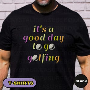 May include: Black t-shirt with the text "it's a good day to go golfing" in colorful letters and golf ball graphics. The shirt also has the word "T-SHIRTS" in a purple box and the word "BLACK" in a black circle.