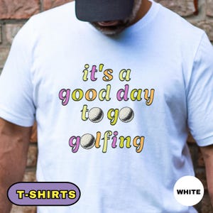 May include: White t-shirt with the text "it's a good day to go golfing" in colorful lettering. The design includes golf balls. The shirt has a crew neck and short sleeves. The words "T-SHIRTS" and "WHITE" are also visible.