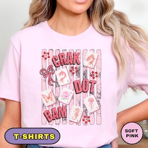 Könnte beinhalten: Ein zartrosa T-Shirt mit einem Grafikdruck aus vertikalen, abgenutzten Streifen, einer rosa Schleife und mehreren Mahjong-Steinen, die mit Illustrationen von Fisch, Schmetterling, Blume, Seepferdchen und Qualle sowie dem Text CRAK, DOT und BAM verziert sind.