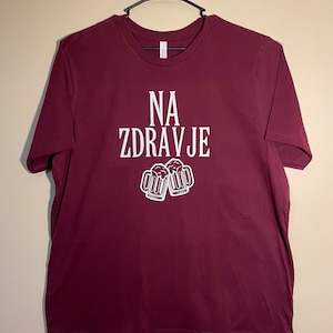 May include: Burgundy t-shirt with the words "NA ZDRAVJE" in white, and a graphic of two beer mugs. The shirt is hanging on a black hanger against a neutral background. A casual wear item.