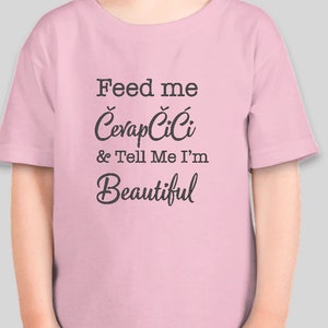 May include: A light pink t-shirt with the text "Feed me Ćevapčići & Tell Me I'm Beautiful" in dark gray lettering. The shirt is short-sleeved and appears to be made of a soft material.