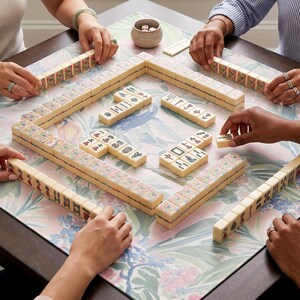 May include: A group of people playing Mahjong on a floral patterned table. The game pieces are cream-colored tiles with various symbols and characters. Dice and small bowls are also visible. The players' hands are visible, arranging the tiles.