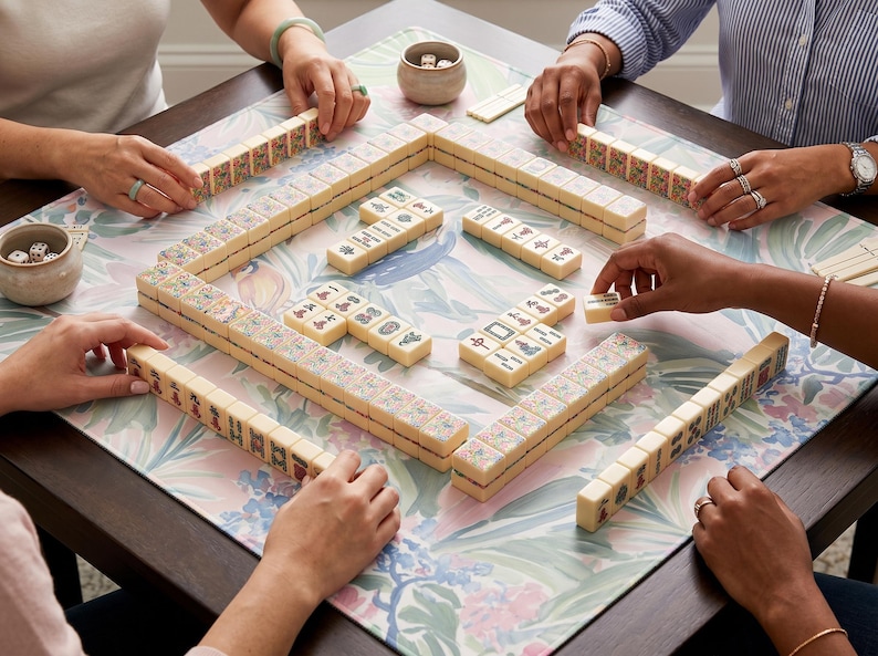 May include: A group of people playing Mahjong on a floral patterned table. The game pieces are cream-colored tiles with various symbols and characters. Dice and small bowls are also visible. The players' hands are visible, arranging the tiles.