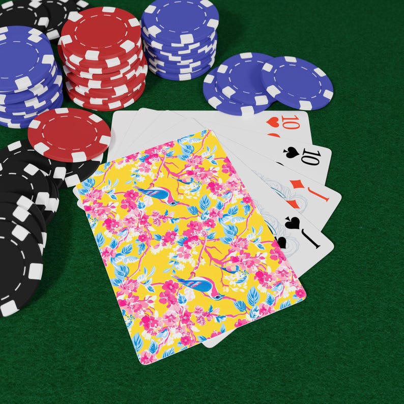 May include: A deck of playing cards with a floral design, featuring pink flowers and blue birds on a yellow background, next to poker chips in red, blue, and black. Four playing cards are visible, showing the number 10.
