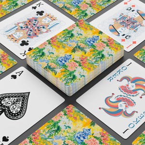 May include: A deck of playing cards with a floral design on the back, featuring yellow, pink, blue, and green colors. The cards are arranged around a deck, with the Ace of Spades, King of Clubs, and Joker cards visible.