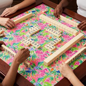 May include: A group of people playing Mahjong on a square table. The table has a dark wood frame and a colorful floral patterned surface with pink, green, and white elements. The ivory-colored tiles are arranged on the table.