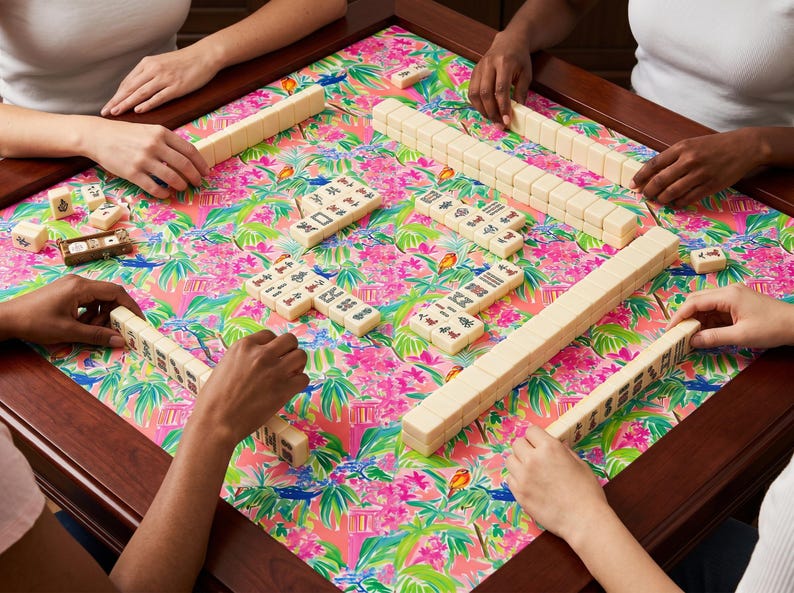 May include: A group of people playing Mahjong on a square table. The table has a dark wood frame and a colorful floral patterned surface with pink, green, and white elements. The ivory-colored tiles are arranged on the table.