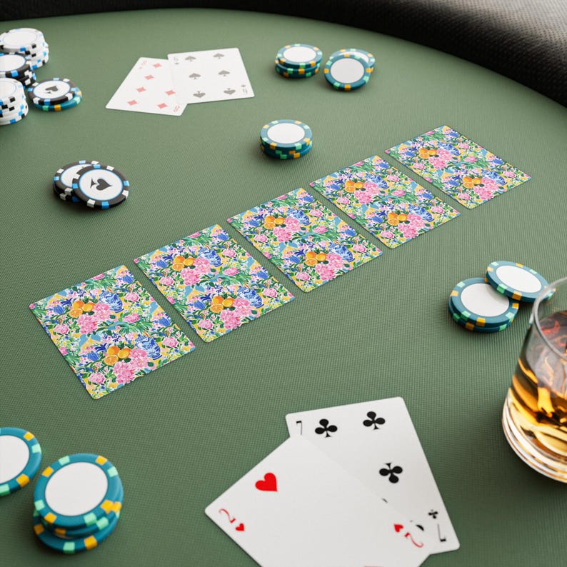 May include: A poker table scene with playing cards and poker chips. The cards feature a floral design with pink, blue, and yellow flowers. Poker chips are in various colors, including white, blue, and green. A glass of amber liquid is visible.
