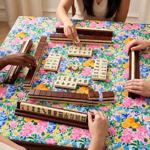 May include: A group of people playing Mahjong on a table covered with a floral patterned cloth. The game pieces are beige with dark brown racks. The table cloth has a colorful pattern of pink flowers, blue vases, and oranges.