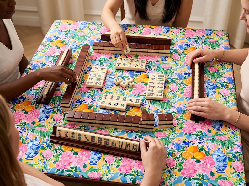 May include: A group of people playing Mahjong on a table covered with a floral patterned cloth. The game pieces are beige with dark brown racks. The table cloth has a colorful pattern of pink flowers, blue vases, and oranges.