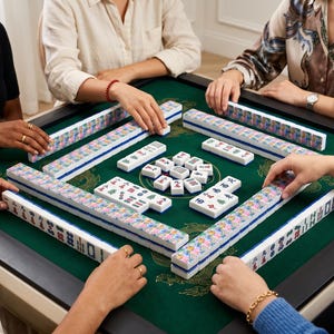 May include: A group of people playing Mahjong. The game is set on a green felt table with white and blue tiles. The tiles are arranged in a square, with players' hands around the edges.