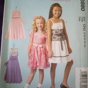 May include: A sewing pattern for a girl's dress. The pattern is for sizes 7-8, 10, 12, and 14. The pattern includes three different styles of dresses: a pink dress with a sash, a white dress with a black lace overlay, and a purple dress with a lace overlay.