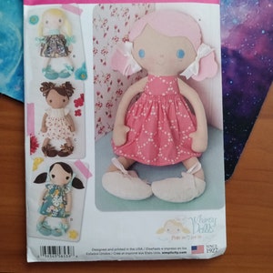 May include: Simplicity sewing pattern for Whimsy Dolls. The pattern features a doll with pink hair and a pink polka dot dress. The pattern also shows three other doll designs. The pattern is printed in the USA.