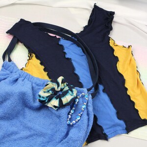 Peut inclure: Un haut sans manches composé de panneaux diagonaux en tissu bleu marine, bleu vif et jaune avec des coutures apparentes à bords ondulés. Un sac en tissu éponge bleu est inclus, orné d'un chouchou tie-dye et d'un bracelet de perles yin-yang.