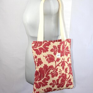 Può includere: Una borsa tote con una base color crema e un audace motivo floreale rosso. La borsa presenta due spallacci color crema e una piccola etichetta rettangolare bianca con testo nero cucita sulla superficie anteriore del tessuto.