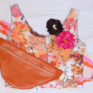 Peut inclure: Un haut sans manches avec un motif floral orange, rose et blanc aux coutures bleues apparentes, un sac banane marron avec une fermeture éclair dorée et deux chouchous en tissu marron foncé et rose vif, le tout disposé ensemble.