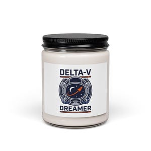 Puede incluir: Un tarro de vela blanco con tapa negra. La etiqueta presenta el texto "DELTA-V DREAMER" con un gráfico de un casco de astronauta, una luna creciente y un cohete en tonos azules y naranjas.