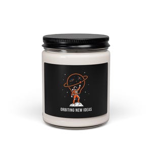 May include: A white candle jar with a black lid. The label features an astronaut in an orange spacesuit holding a planet with an orange ring around it. The text "ORBITING NEW IDEAS" is below the illustration.