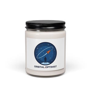 May include: A white candle jar with a black lid. The label features a blue circle with a rocket ship, a person, and the text "ORBITAL OPTIMIST". The candle is a decorative item.