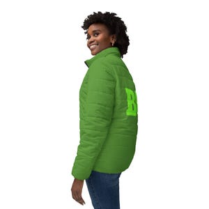 Puede incluir: Chaqueta acolchada verde brillante con cremallera negra y la letra "B" en verde claro en la espalda. La chaqueta se lleva con vaqueros azules.