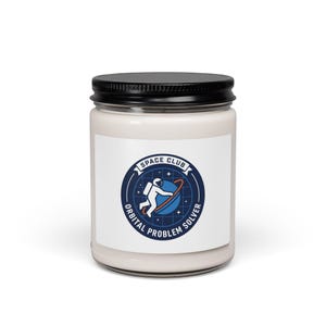 May include: A white candle jar with a black lid. The label features a blue and white graphic of an astronaut orbiting a planet, with the text "SPACE CLUB ORBITAL PROBLEM SOLVER".