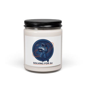 May include: A white candle jar with a black lid. The jar has a label with a blue and orange graphic of an astronaut holding a laptop in space. The text "SOLVING FOR AV" is below the graphic.