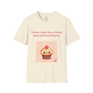 May include: Cream-colored t-shirt featuring the text "Procrasti-baking: The art of baking instead of doing something else." Below the text is a cartoon cupcake with a cherry on top. The cupcake has a smiling face and is decorated with sprinkles.