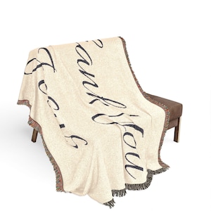 Può includere: Coperta in tessuto color crema con scritta nera "Thank You". La coperta ha un bordo a frange con un mix di colori. La coperta è drappeggiata su una panca marrone.