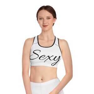 May include: White sports bra with black trim and the word "Sexy" in a cursive font. The bra has a racerback design and is made of a smooth, likely athletic material. The overall aesthetic is clean and modern.