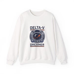 Könnte beinhalten: Weißes Sweatshirt mit Rundhalsausschnitt und einer Grafik eines Astronautenhelms. Das Design enthält den Text "DELTA-V" über dem Helm und "DREAMER" darunter. Die Helm-Grafik zeigt einen Mond, Sterne und eine Rakete.