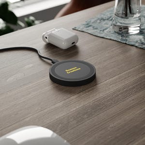 May include: A black wireless charger with the text "Beamon Creations" in yellow, connected to a black cable. A white charging case for earbuds is on a wooden surface.