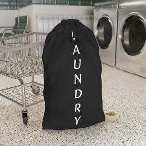 May include: A black laundry bag with the word "LAUNDRY" printed vertically in white. The bag is made of a soft material and has a drawstring closure. A metal laundry cart and washing machines are in the background.