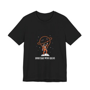 Pode incluir: Camiseta preta com um astronauta de desenho animado em um traje espacial laranja, em pé em uma superfície lunar, alcançando uma lâmpada em forma de planeta com elementos orbitando. O texto "ORBITING NEW IDEAS" está abaixo.
