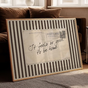May include: Framed artwork featuring a vintage postcard design with the text "It feels so good to be home." The artwork has a striped black and white border and a wooden frame. The piece is displayed in a living room setting.