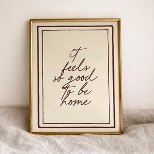 May include: A framed print with the words "It feels so good to be home" in a handwritten style. The print has a cream-colored background with a dark brown border and text, set in a gold-colored frame.