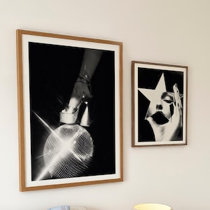 May include: Two framed monochrome art prints. One shows a high-heeled shoe on a disco ball, the other a face with a star cutout. The prints are on a white wall. A lamp and bedside table are in the foreground.