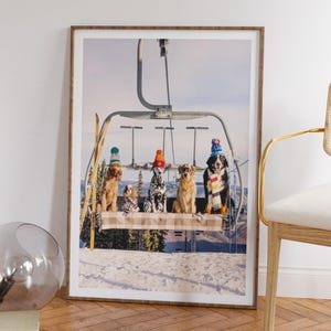 May include: A framed print featuring five dogs wearing winter hats and scarves, seated on a ski lift. The dogs include a Dalmatian, a Golden Retriever, and a Bernese Mountain Dog. The background shows a snowy mountain scene.