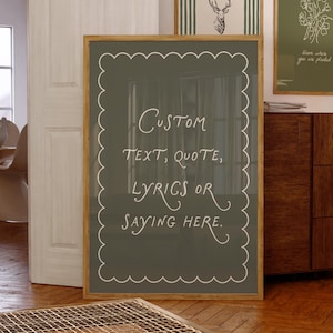 Puede incluir: Una lámina decorativa rectangular en un marco de madera clara con fondo verde oliva oscuro y un borde festoneado blanco. El texto dice: Custom text, quote, lyrics or saying here. La lámina se muestra en un entorno de habitación.