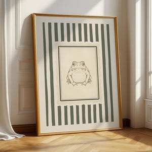 May include: A framed art print featuring a detailed line drawing of a frog. The artwork is set against a cream-colored background with a green and white striped border, encased in a gold frame. The print is leaning against a white wall.