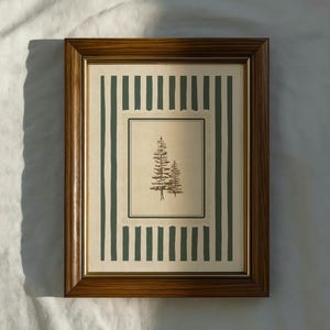 Könnte beinhalten: Ein gerahmter Druck mit einer detaillierten Illustration von zwei Kiefern. Das Kunstwerk befindet sich vor einem cremefarbenen Hintergrund mit einem grünen Streifenmuster. Der Rahmen ist aus dunklem Holz.