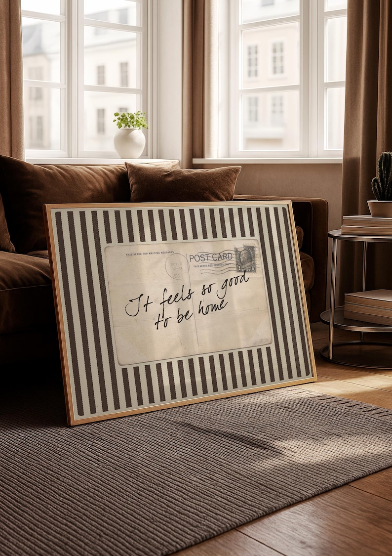 Vintage postcard-style wall art with handwritten script reading “It feels so good to be home,” set on a neutral striped background in a cozy sunlit interior.