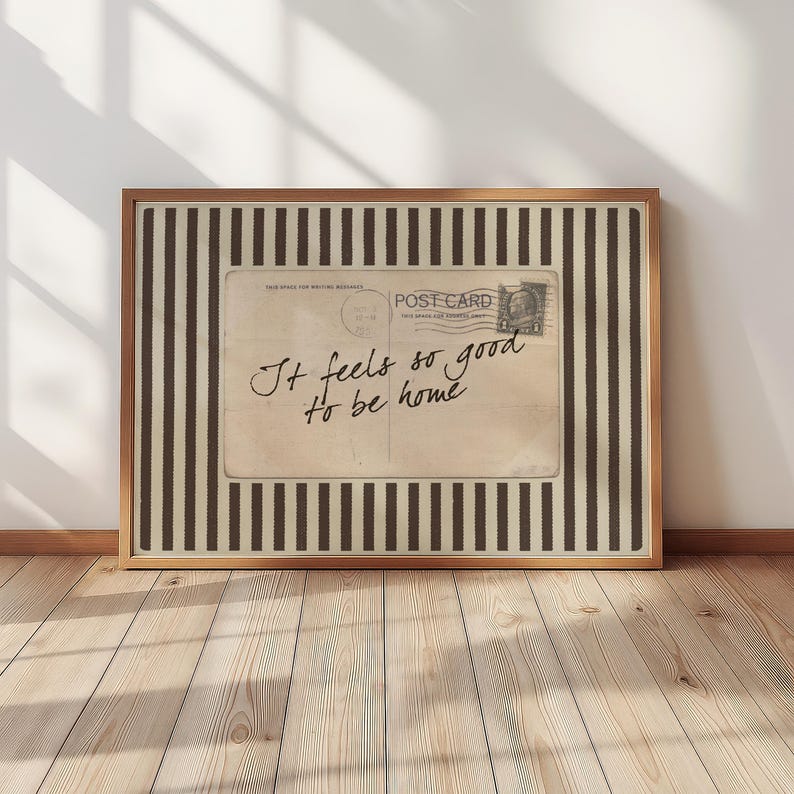 Vintage postcard-style wall art with handwritten script reading “It feels so good to be home,” set on a neutral striped background in a cozy sunlit interior.
